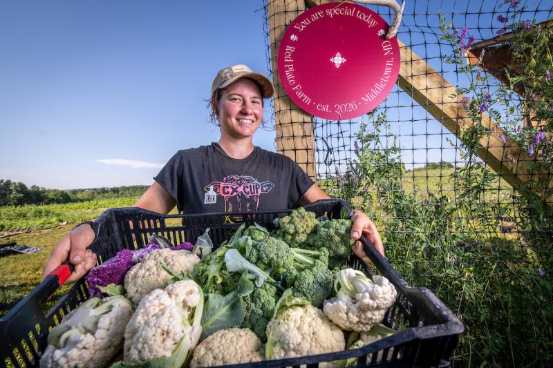 This is a photo of a female farmer holding out a variety of vegetables in a basket